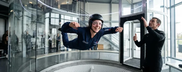 Personne en combinaison de vol flottant dans le tube vitré d'une soufflerie, encadrée par un instructeur debout à l'entrée du tunnel.